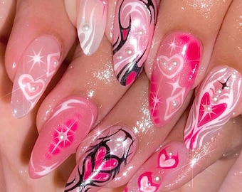 May include: Pink and white almond-shaped nails with a heart and swirl design. The nails are decorated with small white pearls.