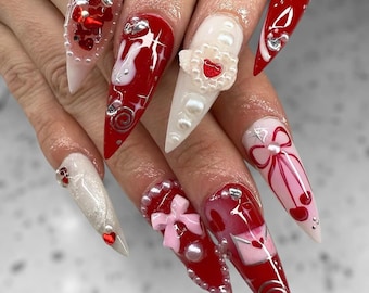 May include: A set of long, pointed acrylic nails with a Valentine's Day theme. The nails feature red, white, and pink designs, including hearts, bows, pearls, and silver accents. Some nails have 3D embellishments.