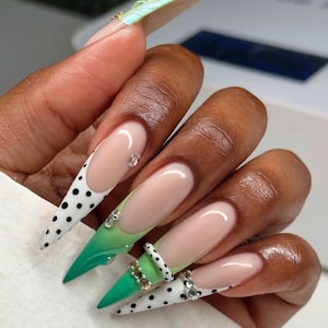 Op de afbeelding: Lange, puntige acrylnagels met een French manicure-ontwerp. De nagels hebben een nude basis met witte punten, zwarte stippen en een groene gradiënt. Sommige nagels hebben strass accenten.