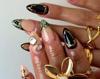 Puede incluir: Primer plano de una mano con uñas largas y almendradas pintadas con un diseño negro y verde. Las uñas están adornadas con detalles dorados y adornos perlados. La mano lleva varios anillos de oro, incluyendo un anillo grande con forma de flor con una perla en el centro.