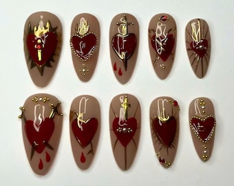 May include: Set of ten false nails with a beige base and red heart designs. Each nail features a different gold design, including a crown, a flame, a sword, and a cross.