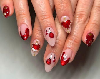 May include: Close-up of a hand with red and gold Christmas-themed nail art. The nails are painted red and have various designs, including hearts, ornaments, and glitter.