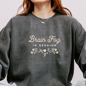 May include: A charcoal gray crewneck sweatshirt featuring the cream-colored text Brain Fog IN SESSION with a decorative floral and heart graphic underneath. The garment has long sleeves and a ribbed neckline, showcasing a washed-out fabric finish.
