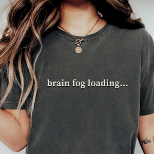 May include: A charcoal gray short-sleeve t-shirt featuring the white text "brain fog loading..." printed across the center of the chest. The garment has a crew neckline and a relaxed fit, showcasing a solid, muted color throughout the fabric.