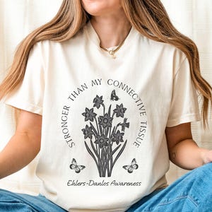 May include: Ivory-colored t-shirt with a round graphic featuring daffodils and butterflies. The text reads "STRONGER THAN MY CONNECTIVE TISSUE" and "Ehlers-Danlos Awareness". The shirt is worn with blue jeans.