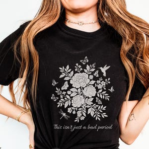 May include: Black t-shirt featuring a floral design with butterflies and a hummingbird. The design includes the text "This isn't just a bad period" in a script font. The shirt is a crew neck.