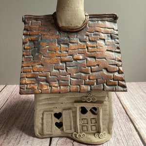 May include: A ceramic cottage with a brown brick roof, a white chimney, and a door with heart-shaped cutouts. The cottage is decorated with small circles and has a rustic, handmade look.