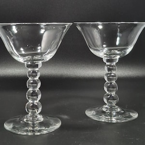 May include: Two clear champagne coupe glasses. Each glass features a wide, shallow bowl and a stem composed of stacked, spherical beads. The glasses sit on a round base, perfect for serving champagne or cocktails.