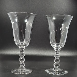 May include: Two clear wine glasses with a flared bowl and a stem featuring a series of small, round beads. The glasses sit on a round base. The background is a solid, dark color.