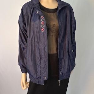 May include: A navy blue windbreaker jacket with a high collar, long sleeves, and elastic cuffs. The jacket features a front zipper closure with snap buttons and a small embroidered diamond pattern in red and white on the upper right chest area.