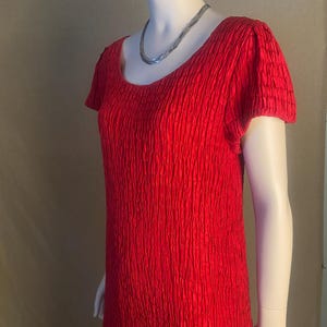 Puede incluir: Vestido rojo brillante, texturizado, con mangas cortas y cuello redondo. El vestido se muestra en un maniquí y se combina con un collar plateado. La tela tiene un patrón de costillas verticales.