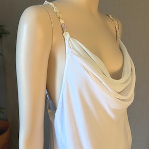 May include: A white, flowing dress with a cowl neckline and delicate floral embellishments on the straps. The dress has a simple, elegant design, perfect for special occasions. The fabric appears soft and drapes gracefully.