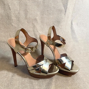 May include: A pair of gold high-heeled sandals. The sandals have a crisscross design over the toes, ankle straps with buckles, and wooden-look heels. The platform heels are approximately 1 inch high, and the stiletto heels are approximately 4 inches high.