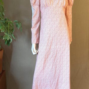 May include: A vintage pink floral print dress with long sleeves and a high neckline. The dress features a white lace panel across the chest and a flowing, floor-length skirt. The dress is displayed on a mannequin.