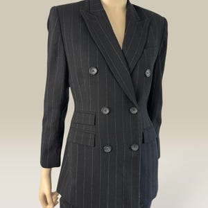 Könnte beinhalten: Ein schwarzer zweireihiger Blazer mit dünnen weißen Nadelstreifen, einem fallenden Revers und sechs dunklen Knöpfen in zwei vertikalen Reihen. Die Jacke verfügt über zwei Pattentaschen auf Taillenhöhe sowie lange Ärmel und ist auf einer Schneiderpuppe präsentiert.