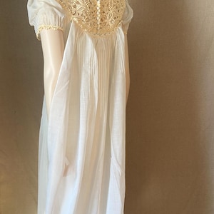 May include: A white vintage nightgown with a cream-colored lace yoke and short sleeves. The nightgown features vertical pleats down the front and small buttons at the back of the neck. The fabric appears to be lightweight and flowing.