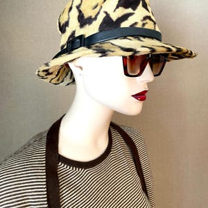 以下が含まれることがあります： 黒い革のバンドとバックルが付いたヒョウ柄の帽子。 サングラスとストライプのシャツを着たマネキンが帽子をかぶっています。 帽子のつばはわずかに湾曲しています。