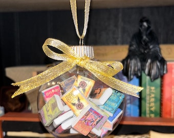 May include: Clear glass Christmas ornament filled with miniature Harry Potter book replicas. The ornament is suspended by a gold ribbon and bow. The background includes a shelf with books and a dark figure.