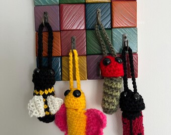 May include: Five crocheted insect keychains hanging on hooks attached to a colourful wooden wall organiser. The insects are a bee, a butterfly, a caterpillar, a ladybug, and a beetle.