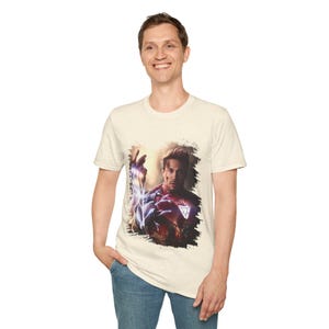 May include: A beige t-shirt with a graphic print of Iron Man in his red and gold suit, with a blue and purple energy effect around his arm. The graphic is set against a black and white background.