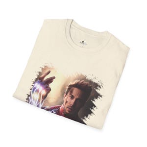 May include: A cream colored t-shirt with a graphic of a man with a determined expression, wearing a red and gold suit, with his hand raised, glowing with energy. The graphic is surrounded by a distressed, faded, brown and white background.