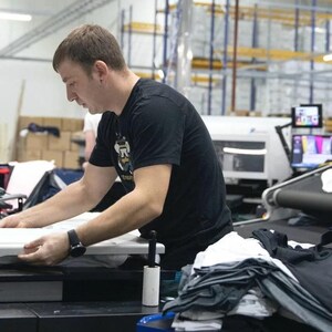 May include: A person working on a large industrial printing machine with a white t-shirt on the conveyor belt. The machine has a black and white control panel with the word "DUAL" printed on it.