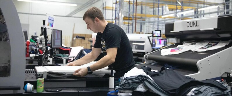 May include: A person working on a large industrial printing machine with a white t-shirt on the conveyor belt. The machine has a black and white control panel with the word "DUAL" printed on it.
