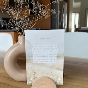 May include: A card with the text "John 14.6-7" and a Bible passage, held in a wooden stand. The card has a watercolour landscape design in beige and green. A beige vase with dried flowers is in the background, creating a serene atmosphere.