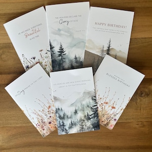 Puede incluir: Una colección de seis tarjetas de felicitación con varios diseños de paisajes en acuarela y citas inspiradoras. Las tarjetas presentan frases como "Beautiful in His Time" y "Happy Birthday!" sobre una superficie de madera.