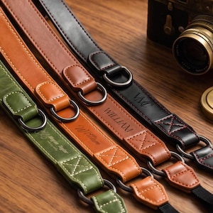 May include: Three camera straps in green, tan, and black leather, each featuring metal D-rings, reinforced stitching, and personalised embossed names. The straps are arranged diagonally, with a vintage-style camera visible in the upper right corner.