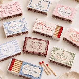 May include: A collection of rectangular matchboxes featuring various decorative designs, including stripes, floral patterns, and text such as Happily Ever After, The Perfect Match, and Cheers To Love, with some boxes open to reveal wooden matches.