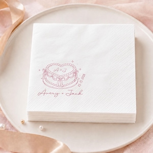 May include: A square white paper napkin featuring a pink line-art illustration of a heart-shaped cake with bows. The text reads A + J, 03.13.2026, and Avery + Jack. The napkin has a diagonal embossed texture pattern along the right side edge.