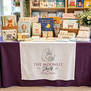 May include: A purple tablecloth featuring a white runner with the text THE MOONLIT Shelf BOOK NOOK and a crescent moon illustration. The table is filled with various children's books, small card sets, and a cup of coloured pencils in baskets.