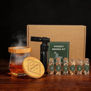 Op de afbeelding: Een whiskey smoker kit met een glazen rookkap, een houten deksel met aangepaste tekst, een brander en kleine glazen flesjes met houtsnippers. De kit bevat een doos en een boekje met de tekst "Whiskey Smoker Kit".