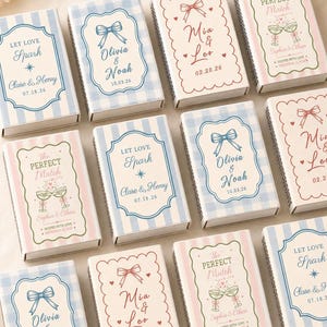 May include: A collection of rectangular matchboxes featuring three distinct designs: blue striped with 'Let Love Spark', blue gingham with a bow and 'Olivia & Noah', and cream with a scalloped border, bow, and 'Mia & Leo' or 'The Perfect Match'.
