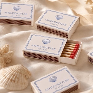May include: Several rectangular matchboxes with white covers featuring a blue scalloped border, a seashell icon, and the text ASHLEY&TYLER 07.19.26. One box is partially open, revealing wooden matches with red tips. The boxes have brown strike strips.