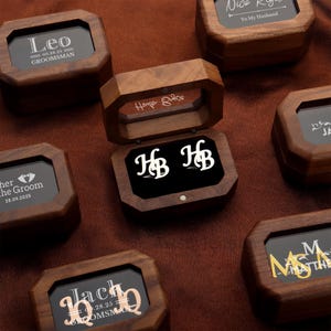 Op de afbeelding: Houten dozen met gepersonaliseerde tekst en data, ideaal als cadeau. Eén doos is open en onthult zilveren manchetknopen met de initialen "HB". Andere dozen tonen namen zoals "Leo" en "Jack", met data en titels.