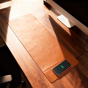 May include: A rectangular, tan leather desk mat with a built-in wireless charging pad. A black smartphone with a charging icon rests on the pad. The mat is on a wooden desk, with the initials "SHM" embossed on the lower right corner.