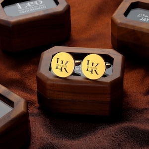 May include: Gold-colored cufflinks with the initials "IV" and the date "06.18.2025" engraved on them. The cufflinks are displayed in a wooden box. Other wooden boxes are visible in the background, one with the word "Leo" on it.