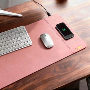 May include: A pink leather desk mat with a wireless charging area, a silver mouse, and a keyboard. A smartphone with a charging symbol is on the mat. The mat has the initials "MOR" in gold.