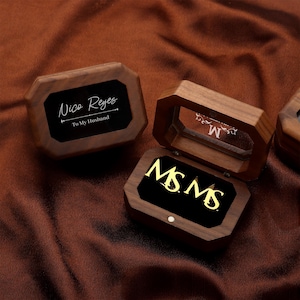 May include: Wooden jewelry boxes with black velvet interiors. One box is open, displaying gold-colored initials "MS MS". Another box lid reads "Nico Reyes To My Husband". The boxes are on a brown fabric surface.