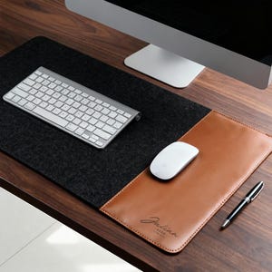 May include: A two-tone desk mat with a black felt section and a brown leather section. A white keyboard, mouse, and a black pen are on the desk. The leather section has the name "Julian Kerr" printed on it.