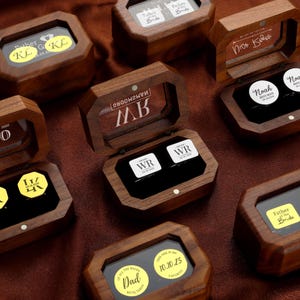 May include: A collection of wooden cufflink boxes, each with a clear lid and personalized details. The boxes are octagonal and contain cufflinks with white surfaces and black text. Some boxes feature phrases like "Father of the Bride" and initials.