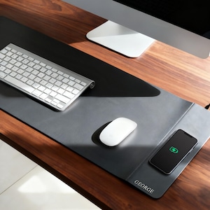 Könnte beinhalten: Eine dunkelgraue Leder-Schreibtischunterlage mit kabelloser Ladestation. Eine silberne Tastatur, eine weiße Maus und ein Smartphone befinden sich auf der Unterlage. Der Name "GEORGE" ist auf die Unterlage gedruckt. Der Schreibtisch ist aus Holz.