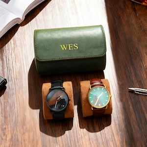May include: A green leather watch case with the initials "WES" in gold lettering. Two watches with leather straps sit on brown watch pillows. One watch has a black band and face, the other has a brown band and a green face.