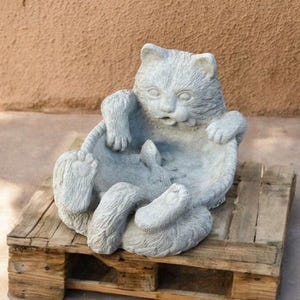 May include: A gray cat statue in a seated position, with its paws and tail visible. The cat is sitting on a wooden pallet. The statue has a surprised expression. The background is a textured brown wall.