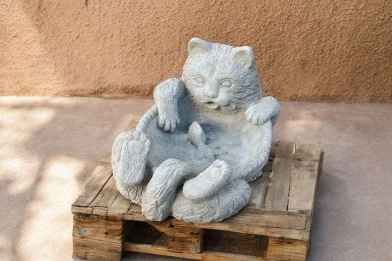 May include: A gray cat statue in a seated position, with its paws and tail visible. The cat is sitting on a wooden pallet. The statue has a surprised expression. The background is a textured brown wall.