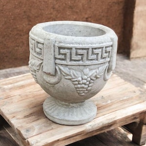 May include: A gray, decorative planter with a pedestal base, Greek key design, and grape cluster motif. The planter is made of a stone-like material and sits on a small wooden stand.