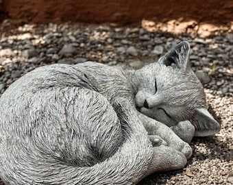 May include: A gray cat statue in a curled sleeping position. The cat's eyes are closed, and its paws are tucked in. The statue is made of a textured material and rests on small pebbles.