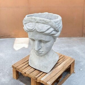 May include: A gray concrete planter shaped like a woman's bust with a simple, round opening at the top. The planter is sitting on a wooden stand.